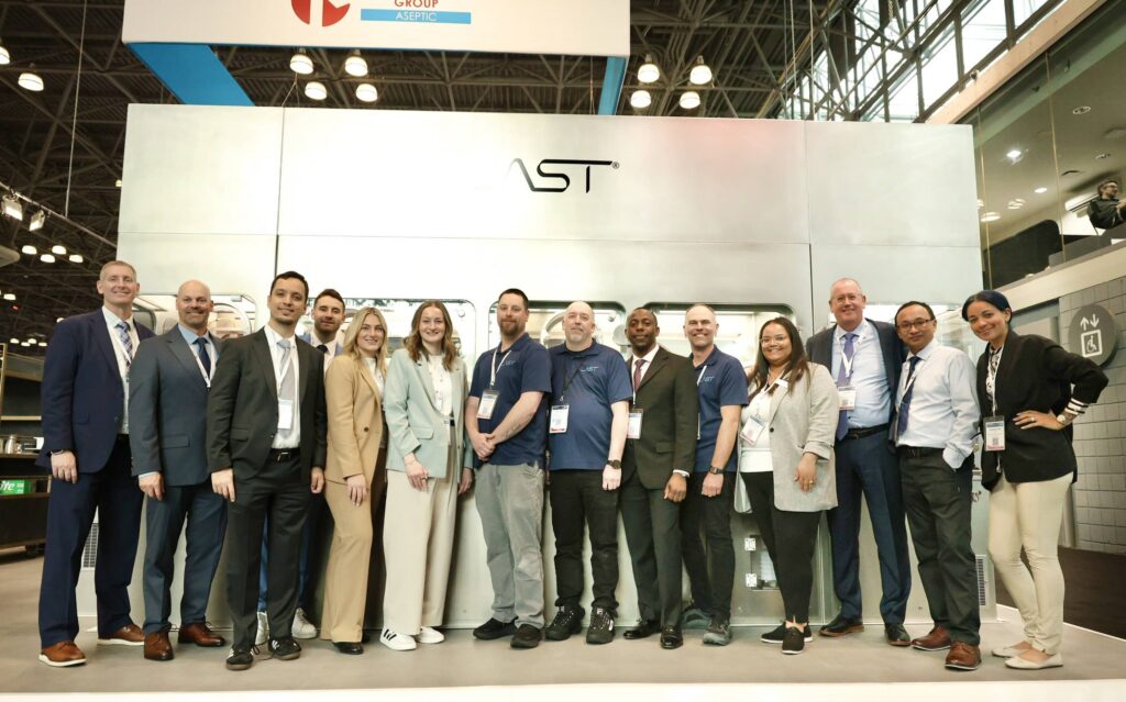 A photo of the AST team that attended INTERPHEX 2026, standing in front of the AST GENiSYS R modular fill-finish system.